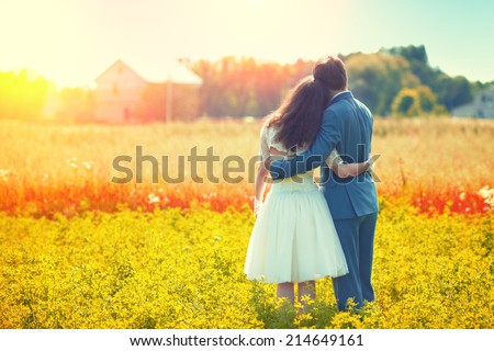 Young happy wedding couple hugging in the meadow back to camera and looking at their house.