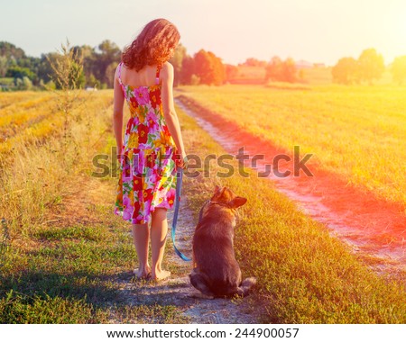 Young woman walking barefoot with dog on rural road back to camera.