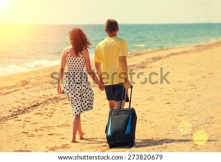 Young couple with travel bag holding hands, walking on the beach back to camera