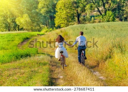 Young happy bride and groom ride bicycles in the meadow back to camera and holding hands