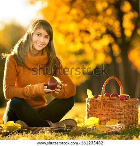 Beautiful young woman is sitting and smiling with rich red apple in her hands and a full basket of tasty rich apples is standing by her on a outdoors autumn sunset background
