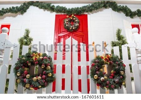 Red front door of the white house nicely decorated for Christmas with fir trees, a wreath and with sparkling lights.