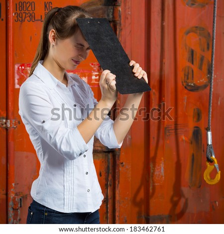 Young business woman on a containers background is hitting herself with a clipboard in despair and anxiety because of a logistics management problem