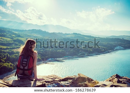 Young tourist smiling woman is sitting on the top of the mounting and with pleasure looking at a beautiful landscape