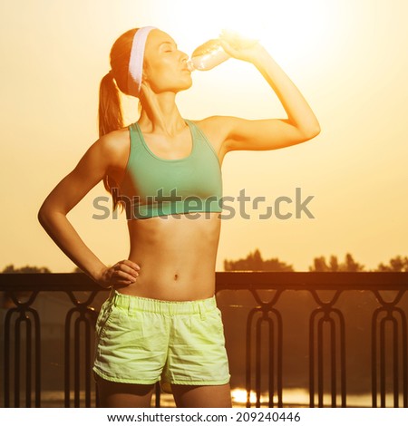Athletic beautiful sports woman is drinking pure water from the bottle refreshing herself after running on a sunny bright light. Healthy sports lifestyle concept.