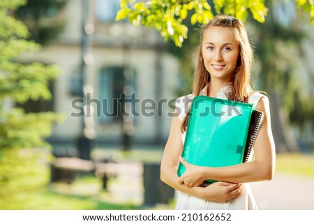 Student woman is standing with the folder and copy-book on the university campus background at the start of a school year in college. Happy smart young woman is going to study outdoors in the park.