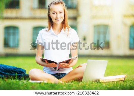 Happy pretty woman is sitting on the lawn in the university campus with notebook in her hands and using laptop. Study outdoors concept.