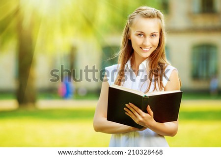Young happy student woman with the book in her hands is standing in smiling in the university campus. Student is studying in the college yard in fall in sunny bright light. Love learning concept.