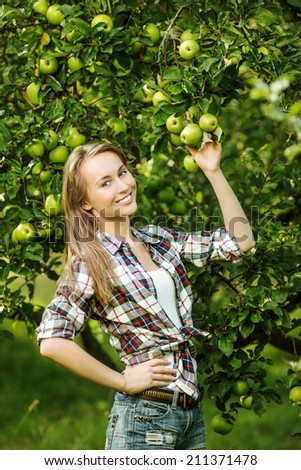 Woman in an apple tree garden during the harvest season. Young smiling beautiful woman is standing among the apple trees with ripe organic apples on it. Healthy country lifestyle concept.