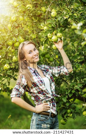 Woman in a sunny apple tree garden during the harvest season. Young smiling beautiful woman is standing among the sunlit apple trees with ripe organic apples on it. Healthy country lifestyle concept.