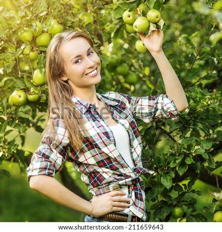 Woman in a sunny apple tree garden during the harvest season. Young smiling beautiful woman is standing among the sunlit apple trees with ripe organic apples on it. Healthy country lifestyle concept.