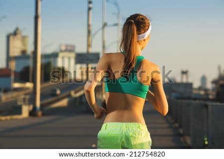 Running woman. Runner is jogging in sunny bright light on sunrise. Female fitness model training outside in the city on a quay. Sport lifestyle. Back view