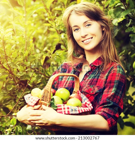 Woman with basket of apples in a garden. Young smiling attractive woman is standing with full basket of organic ripe apples in a sunlit orchard. Country happy lifestyle concept. Harvest season.