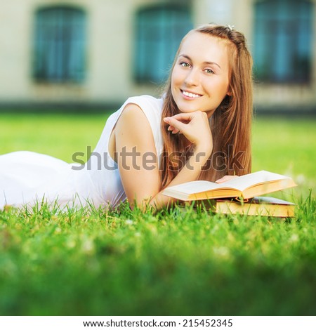 Young happy student woman with the book in her hands is lying and smiling in the university campus on a green lawn. Student is studying in the college yard on a sunny day. Love learning concept.