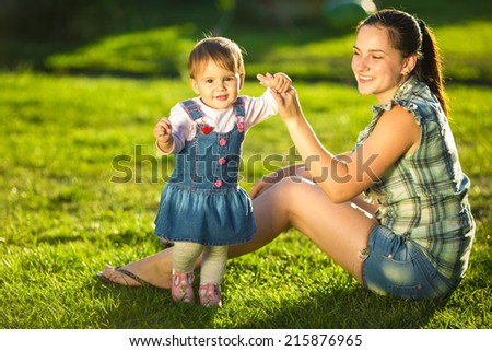 Baby girl is doing her first steps with mothers help. Cute little girl learns to walk with her young mom helping her in the sunny garden outdoors. Happy family  childhood and motherhood concept.