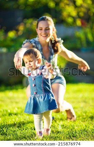 Baby girl is doing her first steps with mothers help. Cute little girl learns to walk with her young mom helping her in the sunny garden outdoors. Happy family childhood and motherhood concept.