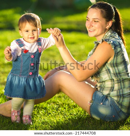 Baby girl is doing her first steps with mothers help. Cute little girl learns to walk with her young mom helping her in the sunny garden outdoors. Happy childhood and motherhood concept.