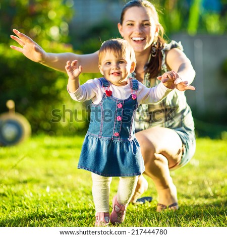 Baby girl is doing her first steps with mothers help. Cute little girl learns to walk with her young mom helping her in the sunny garden outdoors. Happy childhood and motherhood concept.