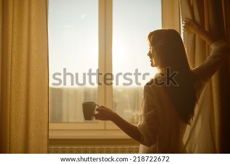 Woman in the morning. Attractive sexy woman with neat body is holding a cup with hot tea or coffee and looking at the sunrise standing near the window in her home and having a perfect cozy morning.
