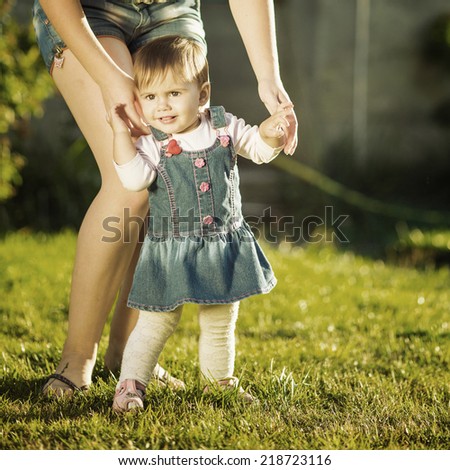 Baby girl is doing her first steps with mothers help. Cute little girl learns to walk with her young mom helping her in the sunny garden outdoors. Happy childhood and motherhood concept. Instagram.