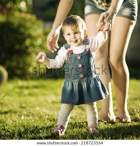 Baby girl is doing her first steps with mothers help. Cute little girl learns to walk with her young mom helping her in the sunny garden outdoors. Happy childhood and motherhood concept. Instagram.