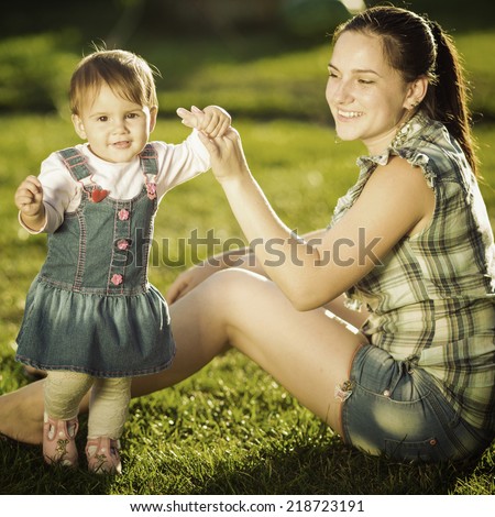Baby girl is doing her first steps with mothers help. Cute little girl learns to walk with her young mom helping her in the sunny garden outdoors. Happy childhood and motherhood concept. Instagram
