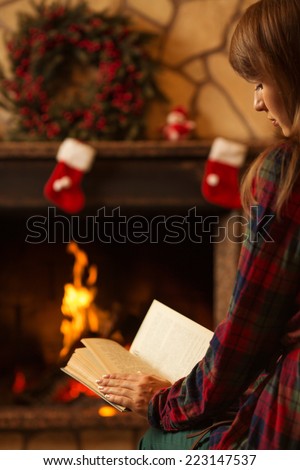 Woman reading a book by the fireplace. Young woman reading a book by the warm fireplace decorated for Christmas. Relaxed holiday evening concept.