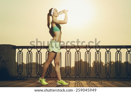 Athletic beautiful sports woman is drinking pure water from the bottle refreshing herself after running on a sunny bright light. Healthy sports lifestyle concept.