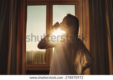 Woman in the morning. Attractive sexy woman with neat body is drinking hot tea or coffee from her cup and looking at the sunrise standing near the window in her home and having a perfect cozy morning.
