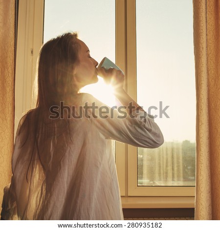 Woman in the morning. Attractive sexy woman with neat body is drinking hot tea or coffee from her cup and looking at the sunrise standing near the window in her home and having a perfect cozy morning.
