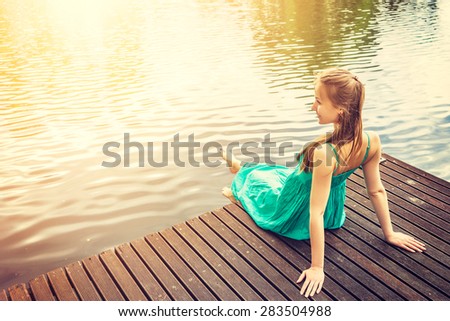 Young woman sitting on a deck by the water, looking into the distance. Carefree bright future concept. Beautiful woman on wooden pier by the lake outdoors. Lens flare sunlight, filter.