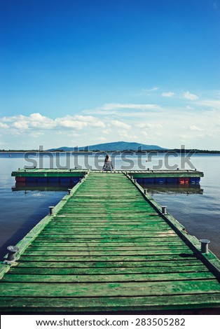 Young woman sitting on a deck by the water, looking into the distance. Carefree bright future concept. Beautiful woman on wooden pier by the lake outdoors on a sunny day.
