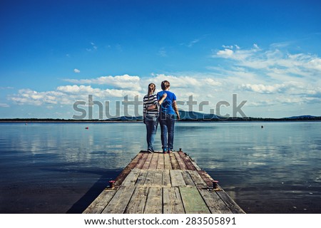 Young couple standing on a deck by the water, looking into the distance. Carefree bright future concept. Hugging man and woman in love on wooden pier by the lake outdoors on a sunny day.