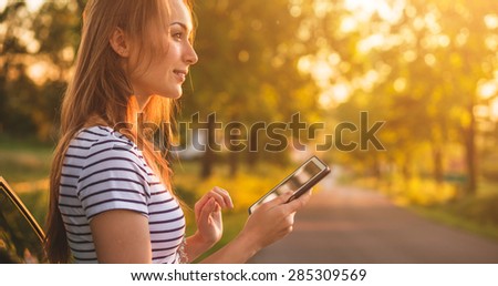 Traveler woman using tablet on car road trip vacation travel. Young woman reading guide book map on tablet computer pc, using mobile internet, relaxing by the car with sunlit road in background.