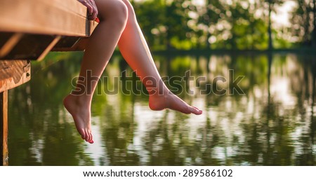 Woman relaxes by the lake sitting on the edge of a wooden jetty , swing one\'s feet near the water surface. Sunny joyful summer day or evening concept.