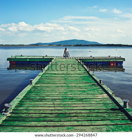 Woman sitting on a wooden jetty enjoying the sunshine with arms up. Woman on a pier on the lake greeting the sun with arms wide open.