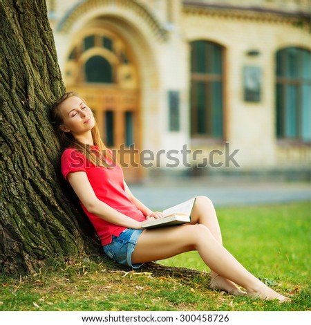 Woman dreaming by the tree with a book. Pretty student girl is sitting near the tree with a book relaxing with joyful and relaxed expression in the university campus. Summer days concept.