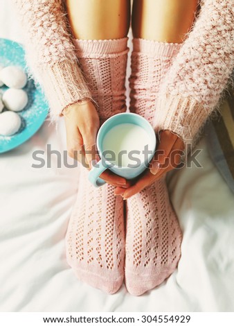 Woman relaxing at cozy home atmosphere on the bed. Young woman with cup of milk in hands and cookies enjoying comfort. Soft light and comfy lifestyle concept.