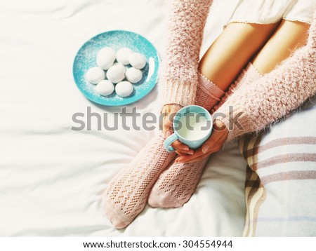 Woman relaxing at cozy home atmosphere on the bed. Young woman with cup of milk in hands and cookies enjoying comfort. Soft light and comfy lifestyle concept.