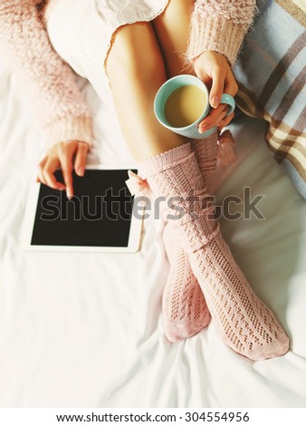 Woman using tablet at cozy home atmosphere on the bed. Young beautiful woman enjoying free time using technological device, holding a cup of cocoa or coffee. Soft light