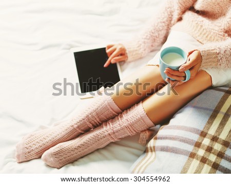 Woman using tablet at cozy home atmosphere on the bed. Young woman with cup of milk in hands enjoying free time with comfort. Soft light and comfy lifestyle concept. Technology in life.