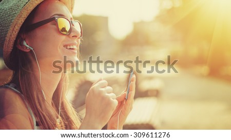 Attractive young woman listening to music on the music player in the city outdoors. Hipster girl enjoying the tunes in her earphones in the morning park. Lens Flare.