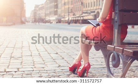 Woman in red high-heeled shoes and dress using digital tablet. Beautiful sexy business woman enjoying sunny morning in the city, sitting on the bench, browsing Internet and drinking coffee. Lens Flare