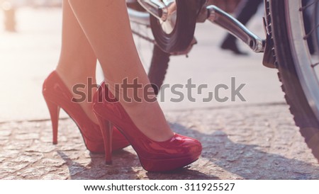 Business woman taking a bicycle form a city-bike station. Close up on feet. Sexy female legs in red high-heeled shoes approaching bicycle and walking away with it. Lens Flare.