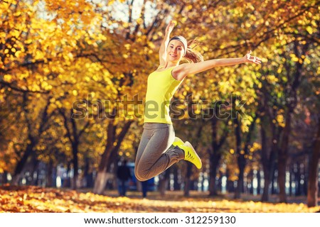 Young happy woman jumping in autumn park. Female fitness model training outside on a warm fall day. Sport lifestyle.