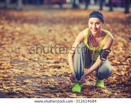 Running woman. Runner is tying laces and listening to music. Female fitness model tying laces outside in autumn park. Woman athlete training in the fall outdoors background. Sport lifestyle.