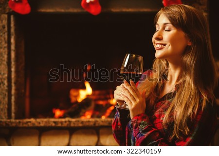 Woman with a glass of wine by the fireplace. Young attractive woman sitting by the fireside and holding a wineglass, enjoying cozy evening. Holiday time concept in a house decorated for Christmas.
