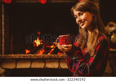 Woman with a mug by the fireplace. Young attractive woman sitting by the fireside and holding a cup with hot drink, enjoying cozy evening. Holiday time concept in a house decorated for Christmas.