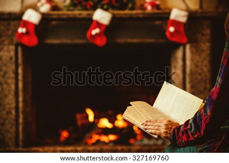 Woman reading a book by the fireplace. Young woman reading a book by the warm fireplace decorated for Christmas. Relaxed holiday evening concept.