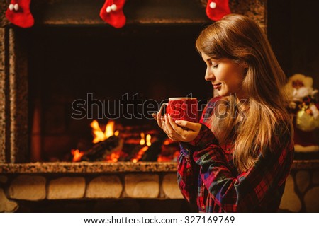 Woman with a mug by the fireplace. Young attractive woman sitting by the fireside and holding a cup with hot drink, enjoying cozy evening. Holiday time concept in a house decorated for Christmas.